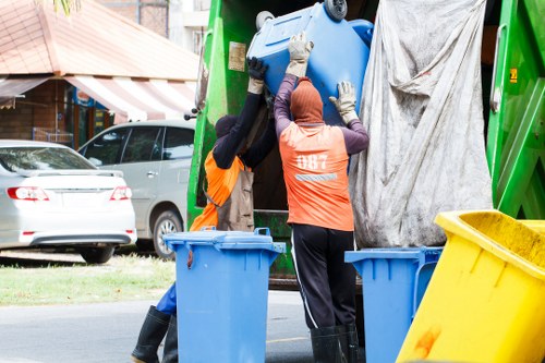 Two waste management workers wearing high-visibility orange vests and gloves are seen loading a large blue plastic bin into a green rubbish collection vehicle on a residential street, with a white car parked on the side and houses with awnings in the background. One worker is supporting the bin while the other guides it into the vehicle's opening. There is also a yellow bin positioned nearby on the pavement. The scene appears to be part of a rubbish collection or clearance operation, possibly associated with office or residential waste disposal services in the Shortlands or nearby area, managed by Office Clearance Shortlands. The environment is outdoors with natural daylight, and the street setting includes trees and typical suburban surroundings, indicating an organized effort to remove rubbish from local properties.