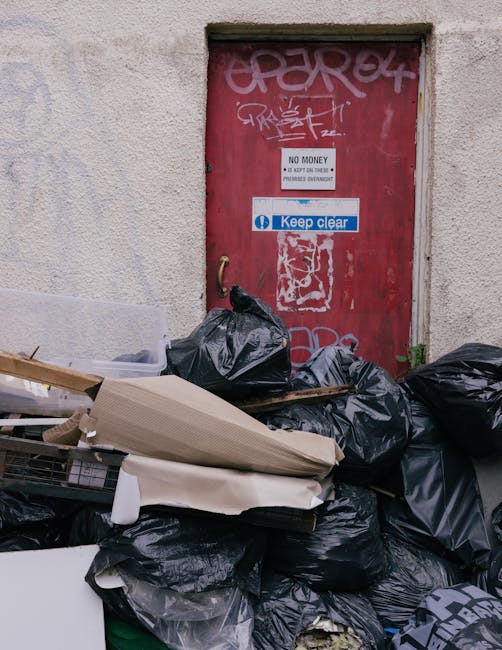A collection of various discarded rubbish bags, primarily black, piled against a beige stucco wall near a red metal door covered in graffiti. The rubbish bags appear full and are stacked haphazardly,