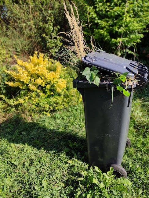 A person dressed in a dark blue work uniform and wearing a wide-brimmed straw hat is crouched down, placing or removing garden waste such as leaves and small branches into a grey plastic rubbish bin. 
